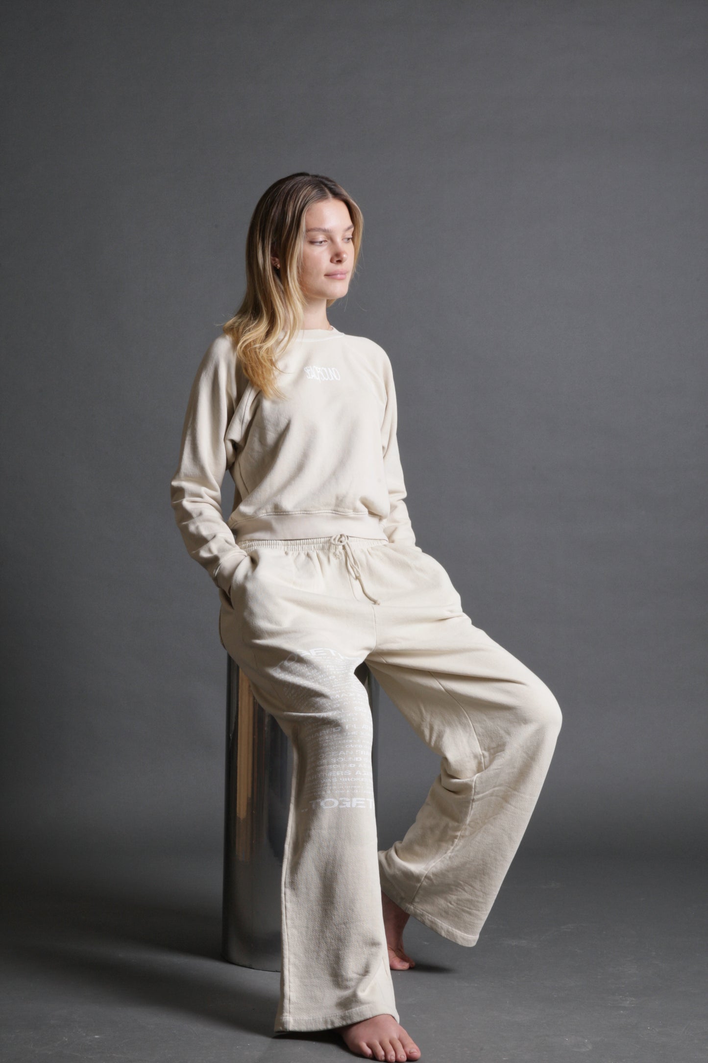 This model is sitting while wearing a crewneck in sand with the Sea Of Sound by Gavin Rossdale Sea Of Sound logo on the back